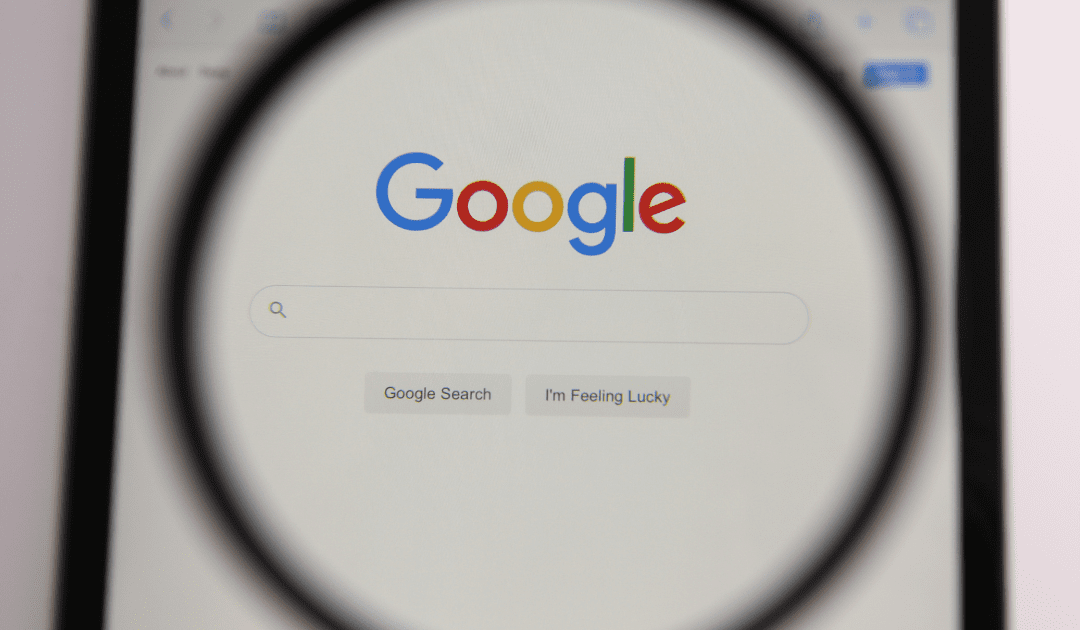 A magnifying glass is positioned over a computer screen displaying the Google homepage. The Google logo is prominently shown above a search bar, with the "Google Search" and "I'm Feeling Lucky" buttons visible below.